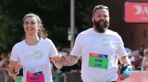 Harriet Davidson Harriet and her husband Adam running the Great Bristol 10k run. They are holding hands as they approach the finish line. They are both wearing white LAM Action charity T-shirts.