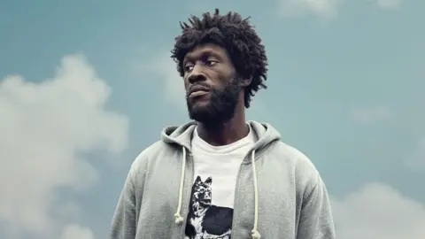 Stormzy as Tenzman in Big Man. He wears a grey hoodie over a white T-shirt with a print of a dog on it. He looks to the left of the camera with a despondent expression and is shot against a pale blue cloudy sky. 