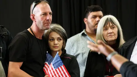 A man and woman embrace while another woman to their left listen to a speaker while holding American flags.