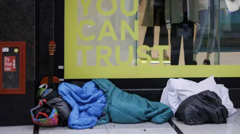 EPA A person sleeping rough, entirely covered in sleeping bags, rests in front of a shop window on Oxford St. There are bags of different colours and sizes at their head and feet and a guitar propped against the window behind them.