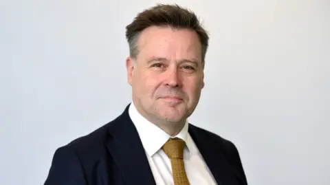 Telford College A man with short dark hair, a dark blue jacket, white shirt and gold tie against a white background