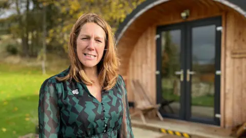 Paula Ward, wearing a green and black dress, outside a holiday pod with trees is the background