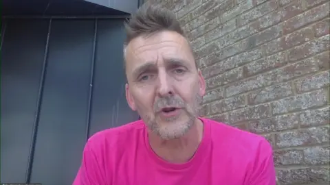 Colin McUke is looking at the camera, has blue eyes and grey hair. He is wearing a bright pink t-shirt.