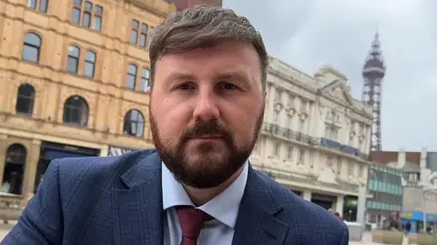 Chris Webb MP with brown hair and beard wearing a navy blue suit and red tie standing outside Blackpool Winter Gardens with Blackpool Tower in the background. 