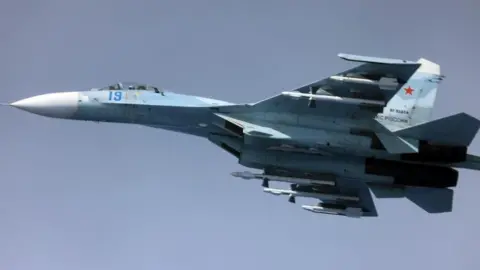 Image of a Russian Su-27, a whit and blue coloured fighter jet, airborne against a clear blue sky.