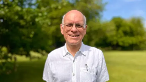Martin Giles/BBC A smiling man faces the camera. He wears glasses, a white shirt with a pocket. A silver coloured pen is visible in the pocket. There is a blurred background of trees and a field
