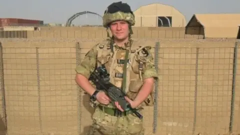 Pete White Pete is wearing beige-khaki army uniform, wearing a helmet with goggles and holding a gun. He's pictured in front of what looks like a beige-coloured aerodrome, and is looking into the camera and smiling slightly.