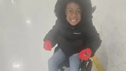 supplied A young boy in a large black coat and Spiderman gloves beams as he sits in a black wheelchair