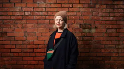 Malachy McCrudden Amberlea is standing in front of a red brick wall. She is looking straight at the camera and is wearing a brown hat. She has short blonde hair and an orange jumper with a navy overcoat. 