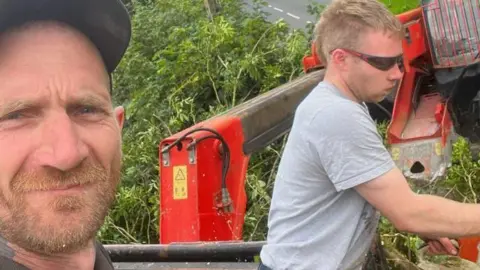 Daniel Graham, wearing a black baseball cap and with a ginger beard, takes a selfie of him and Adam Carruthers, who is wearing sunglasses and a grey shirt, with short fair hair, and is cutting at a tree with a chainsaw