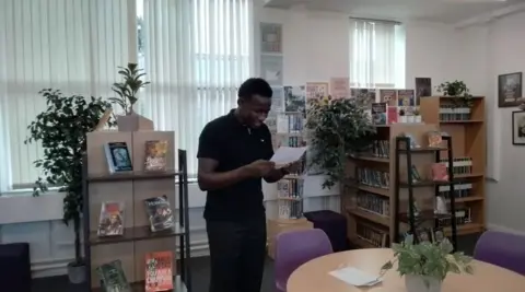 Lift Four Dwellings Secondary Yusuif, wearing a black polo shirt and black jeans in the centre of the image, reads from a piece of paper in a library, surrounded by shelves of books and plants of varying sizes.
