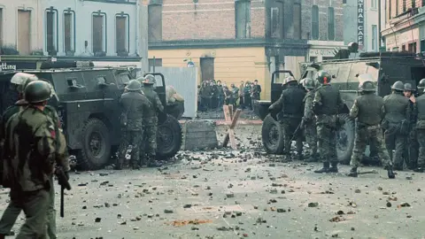 Getty Images Two black army jeeps are parked at a barricade on a street in Londonderry. There are soldiers behind the jeeps taking shelter, they are armed and wearing camouflage clothing and hard hats. In the distance there is a group of young people. There is stones and other debris scattered across the street.