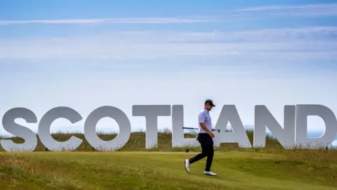 PA Media A lone golfer, photographed side-on, walks along the fairway past a Hollywood-style Scotland sign. A blue sky is visible in the background.
