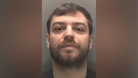 Merseyside Police A police mugshot of Lee Harrison, 27, who has brown hair and a brown beard and smirks at the camera.