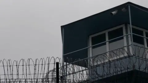 Rayhan Demytrie/ BBC A picture of a grey watchtower with white frames around the windows. Barbed wire is strung along the top of the nearby fence at the bottom of the picture. CCTV cameras can be seen pointing into the prison.