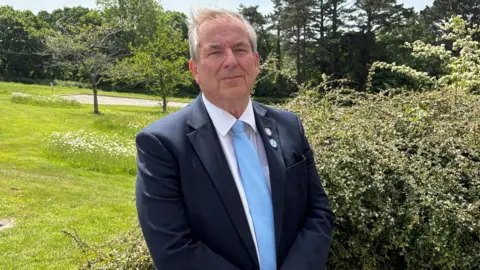 Robert Parsonage is looking directly at the camera as he stands in a garden with lawn, flowers and trees behind him. He has grey hair and is wearing a navy jacket, white shirt and pale blue tie.