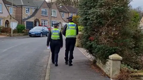 North Yorkshire Police Two officers in police uniform walk along a pavement. Hedges can be seen on the right, while a row of houses can be seen on the left. A blue car is parked in front of one of the houses.