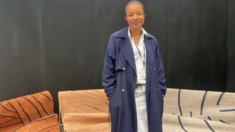 Adaobi Tricia Nwaubani Kelechi Ejikeme in a long navy coat stands in front of jute rugs - mostly brown and beige with navy line designs - at the Decorex show in London