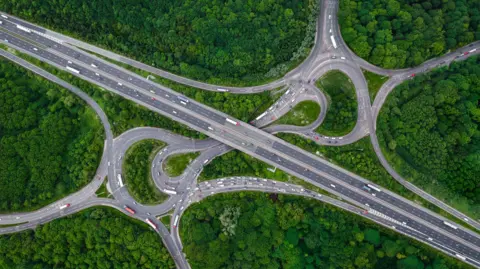 Getty Images Aerial landscape view directly above a busy motorway junction cutting a swathe through dense woodland with green tree canopy cover providing carbon capture from exhaust fumes.