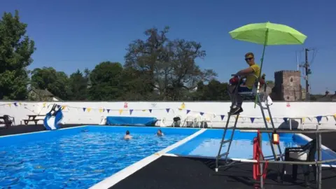 Greystoke Pool A lifeguard on a high chair with a sunshade, watching over two swimmers