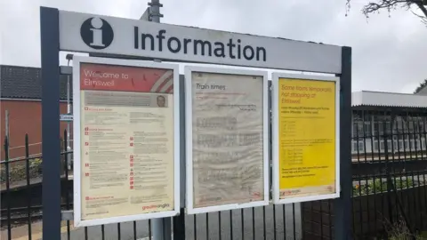 An information board with three separate notices at Elmswell railway station. The platform and railway tracks are behind it.