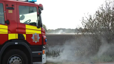 ECFRS A red and yellow fire engine with fields behind it and smoke billowing up