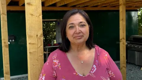 There is a woman with brown shoulder-length hair, a pink flowery blouse and a flower pendant around her neck. She is stood underneath a wooden frame which is used by the snack shack to stay dry. Behind her is the shack, which is painted dark green.