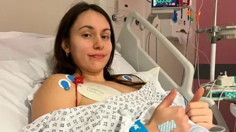 Faith Harrison Faith has long dark hair and is sitting in a hospital bed with a white and blue blanket on. She has stickers on her shoulder, chest and hands. She is smiling and doing a thumbs up motion with two of her hands.