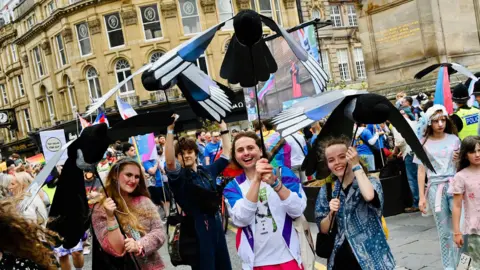 Haydn Brown Four people holding up large magpie puppets above their heads. They are standing in front of Grey's Monument where there is a large crowd of people. The puppet holders are all smiling into the camera. The woman on the left is wearing a pink jumper and has long, blonde hair. To the right is a person with short, dark hair and is wearing a denim jumpsuit. To the right is a man with short, brown hair who is wearing a white and blue sports jacket. On the right is a woman with her brown hair tied back and who is wearing a blue patterned shirt.