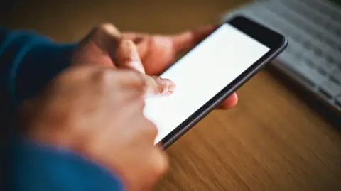 A man's hands tap the blank screen of a cellphone. A computer keyboard can be seen in the background.