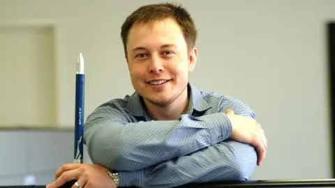 Getty Images Elon Musk shown in 2004 wearing a blue shirt and smiling as he looks at the camera. He is holding a small, blue and silver toy rocket which bears the "SpaceX" logo.