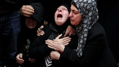 A woman dressed entirely in black is held by another, clutching her hands to chest and her mouth open in a wail.