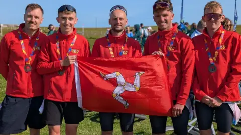 ROB SORBY Members of the men's mountain bike team standing together in a row holding up a Manx flag. They are wearing black shorts and red team Isle of Man tops and have bronze medals hanging around their necks.