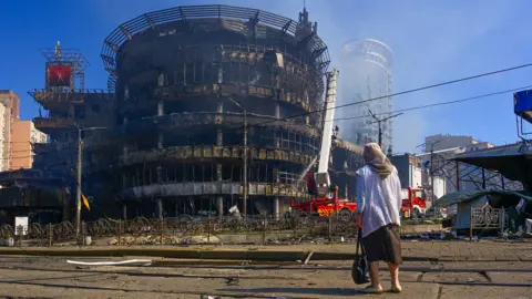A woman seen from behind, wearing a headscarf, watches as firefighters extinguish a fire in a large round building - smoke can be seen rising into the blue sky