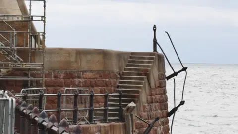 Sunderland City Council Metal railings hanging off Roker Pier. The pier is built from stone and has a dozen or so steps leading up to a higher area. The sea can be seen in the background.