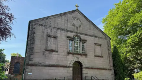 Paul O'Gorman/BBC An old grey brick church is surrounded by large green trees.