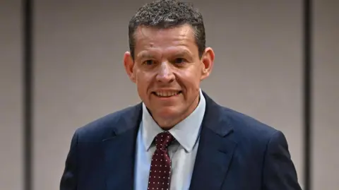 Senedd Cymru Rhun ap Iorwerth looking into a camera, wearing a dark blue suit and a light blue shirt. He is wearing a red tie with white spots.