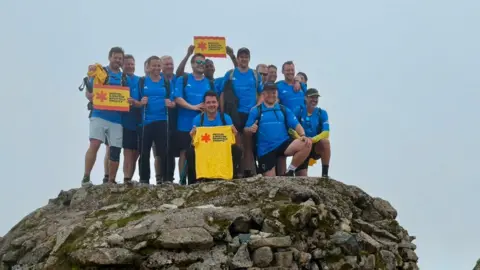 Nick Powell A group of about 15 men stand at the rocky summit of a mountain. They are all wearing blue T-shirts and holding up yellow T-shirts and banners with "Bristol & Weston Hospitals Charity" written on them. They are all smiling and surrounded by a grey sky.