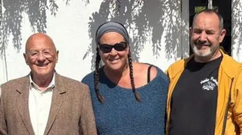 Supplied An older man wearing a brown suit jacket over a white shirt while smiling. He is standing next to a woman wearing sunglasses and a blue jumper and a man wearing a yellow jacket of a black T-shirt whose eyes are closed.