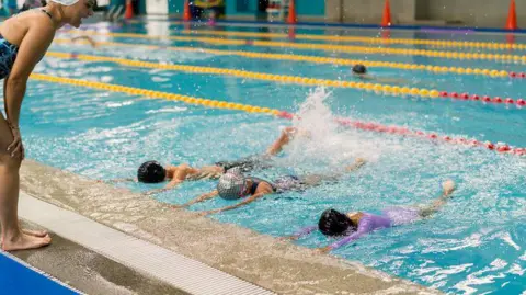 Getty Images Children swim lengths in marked lanes during a lesson at an indoor pool while an instructor stands at the edge watching, with water splashing as they practise.
