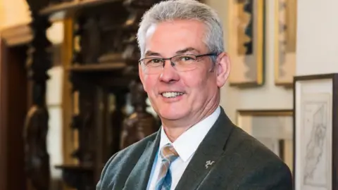 Crookall, who has short grey hair and is wearing glasses, smiling.