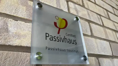 Supplied A close up shot of a brick wall that has a small glass square plaque bolted on with silver knobs. It says Certified Passivhaus / Passivhaus Institut
