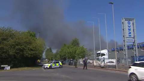 A large black smoke plume can be seen blowing over an IVECO garage with a parked lorry behind a large fence. A police car is parked across the street and a firefighter is walking in the direction of the smoke.