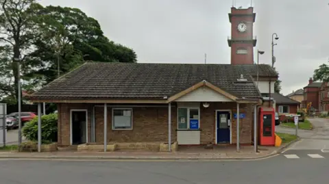 Google A red phone box sits on the right hand side of a small hospital building. A clock tower can be seen in the distance. 