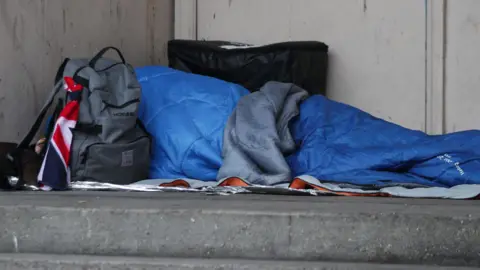 PA Media A homeless person sleeping rough in a doorway. They are lying in a blue sleeping bag with their belongings around them. A grey top and backpack lie besides them.