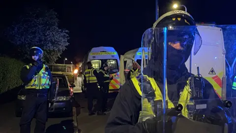 PA Media Police officers wearing riot gear stand in front of police vehicles. Several police vans  can be seen in the background. A blue light glows on the plastic shield one officer holds in front of his face 