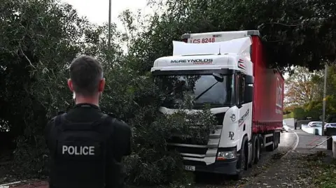 Police officer in black clothing in front of fallen tree and white and red lorry