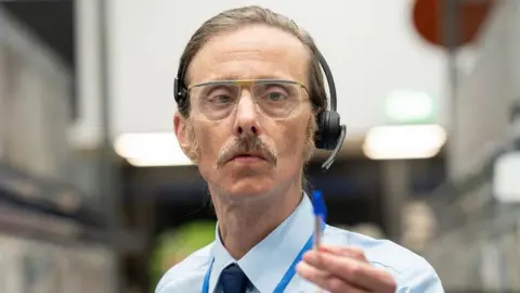 Mackenzie Crook as a shop worker with headset and glasses. he wears a shirt and tie and points with a pen.