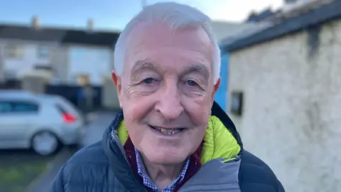 David Burke is outside. he's wearing a navy jacket. The lining is yellow. His shirt is blue and white. 