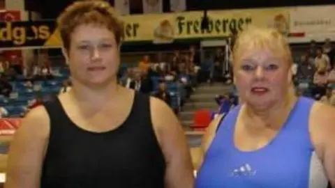 Adele Jones Jones and Bates standing side by side in what appears to be an indoor sports venue or arena. Both are wearing athletic singlets, one in black and the other in blue, along with traditional white sumo belts and competition tags marked “GBR”. They are standing on a carpeted area with rows of spectator seating visible in the background, along with banners and signs hanging around the venue.